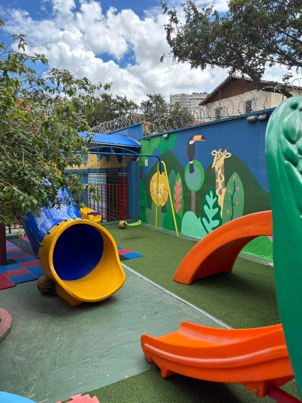 Pátio externo vazio, com escorregadores coloridos, piso emborrachado, muro pintado e árvores ao redor