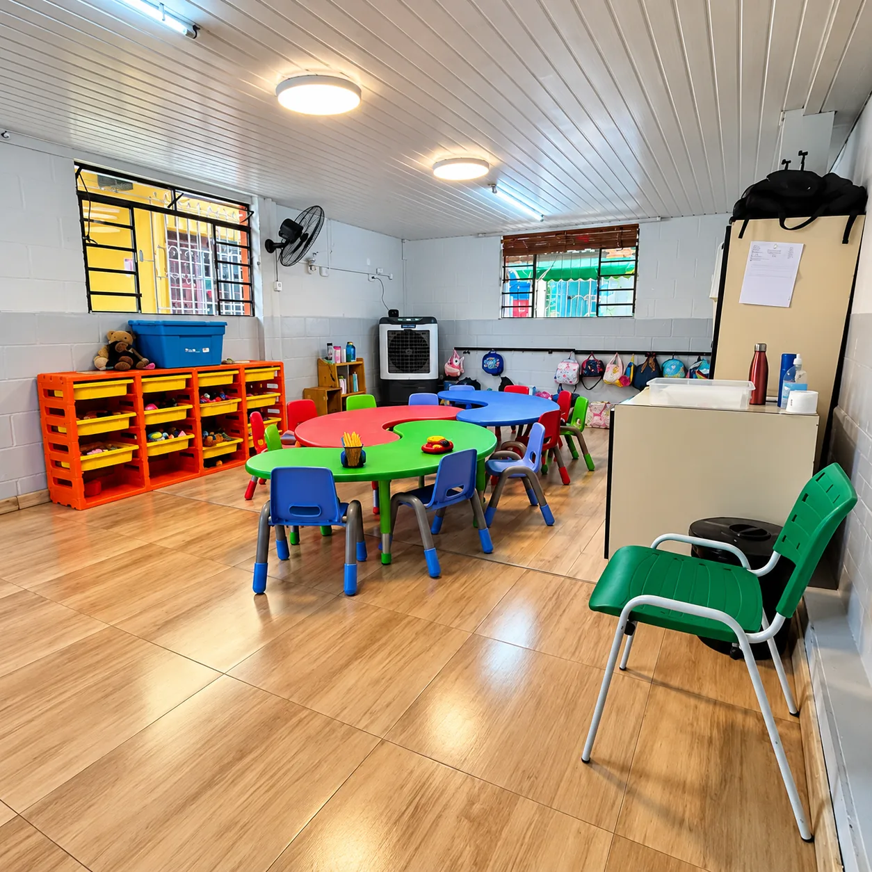 Sala infantil vazia, com mesas e cadeiras coloridas, estantes com brinquedos e janelas gradeadas