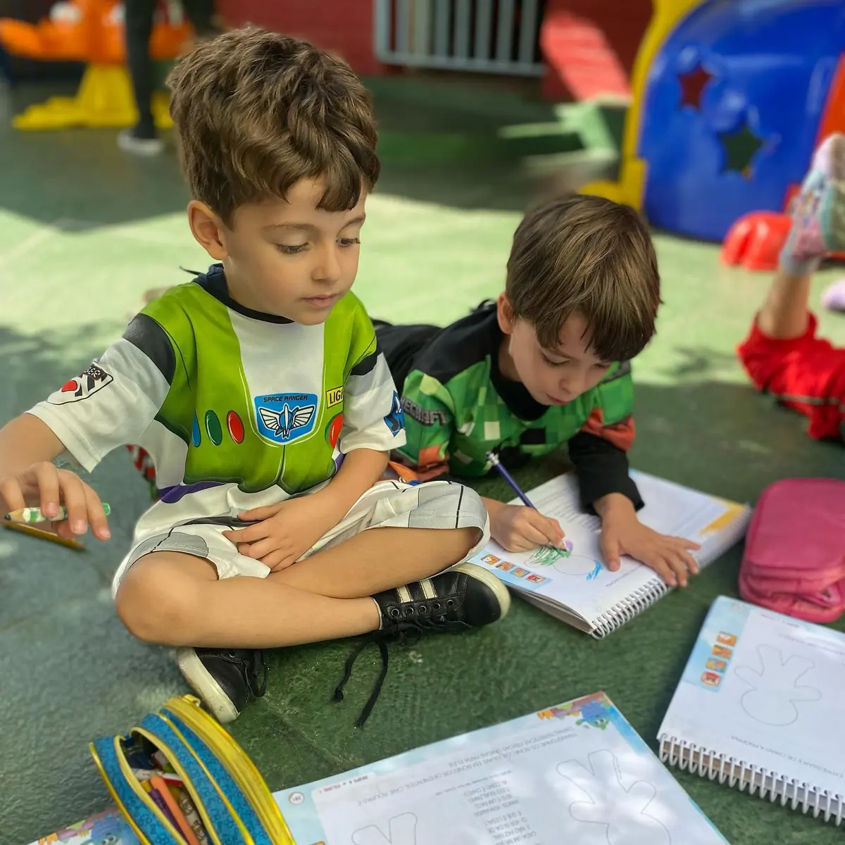 Dois meninos concentrados desenhando lado a lado no chão do pátio, em uma atividade de sala