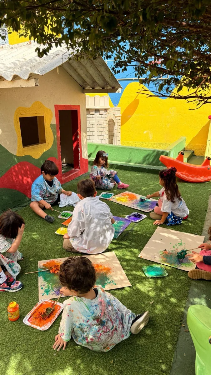 Crianças pintando em folhas sentadas na grama de um pátio, com uma árvore e uma casinha ao fundo