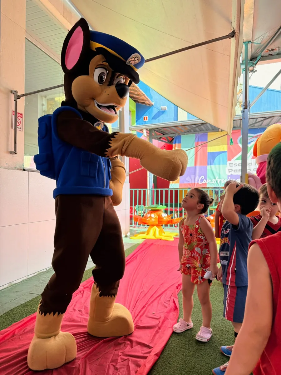 Mascote de cachorro de uniforme azul estendendo a pata para uma menina, com crianças observando em uma área coberta de recreação